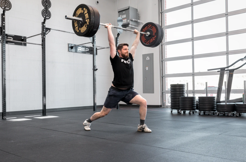 A man performs a clean and jerk using a barbell in a gym.